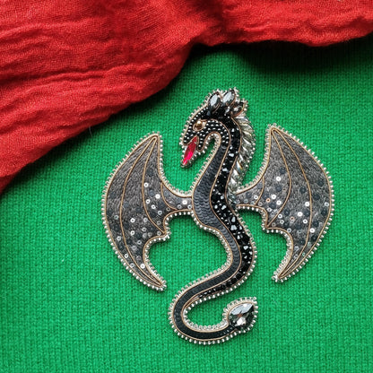 A decorative dragon brooch with a black and silver design, featuring a red eye and wings, is displayed on a green fabric background.