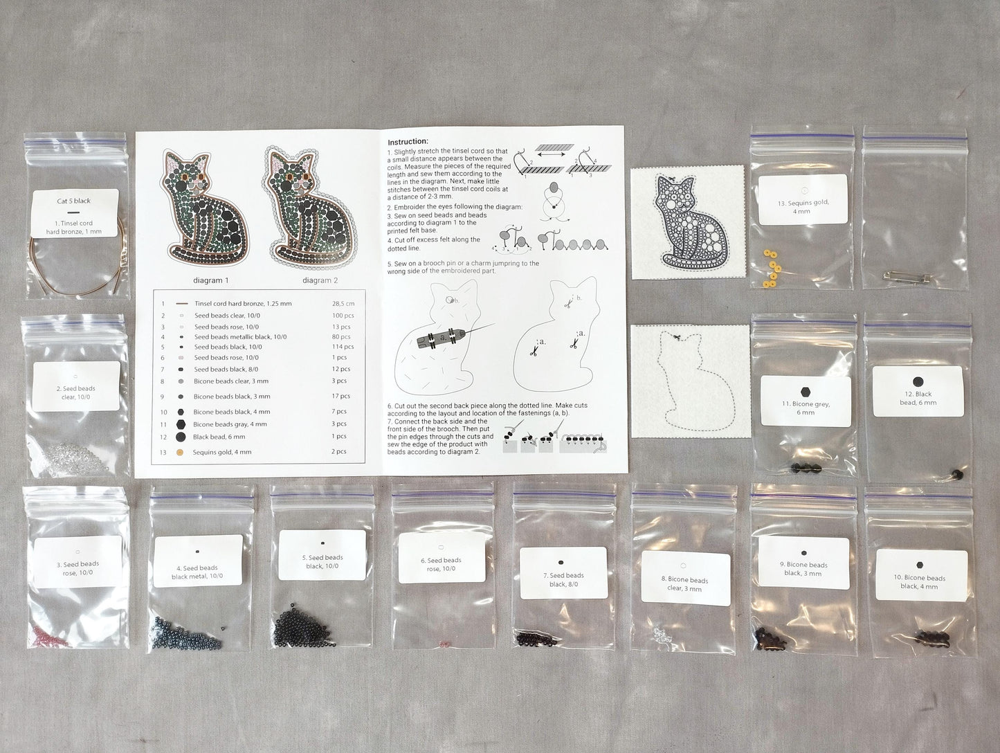 various craft supplies, including bags of beads, seeds, and other materials, arranged on a table. There is also a printed guide or instructions sheet for a project involving cat-themed items, such as earrings and cat-shaped objects.