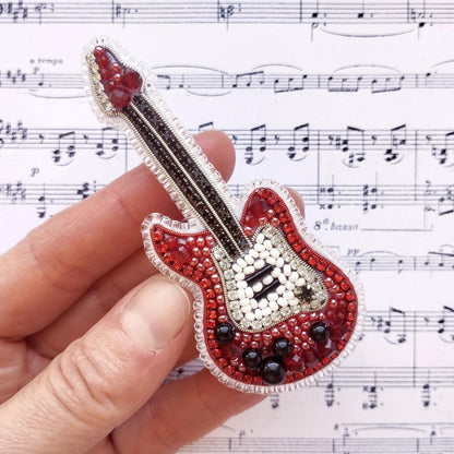A hand holding a red and white beaded guitar brooch against a background of sheet music.