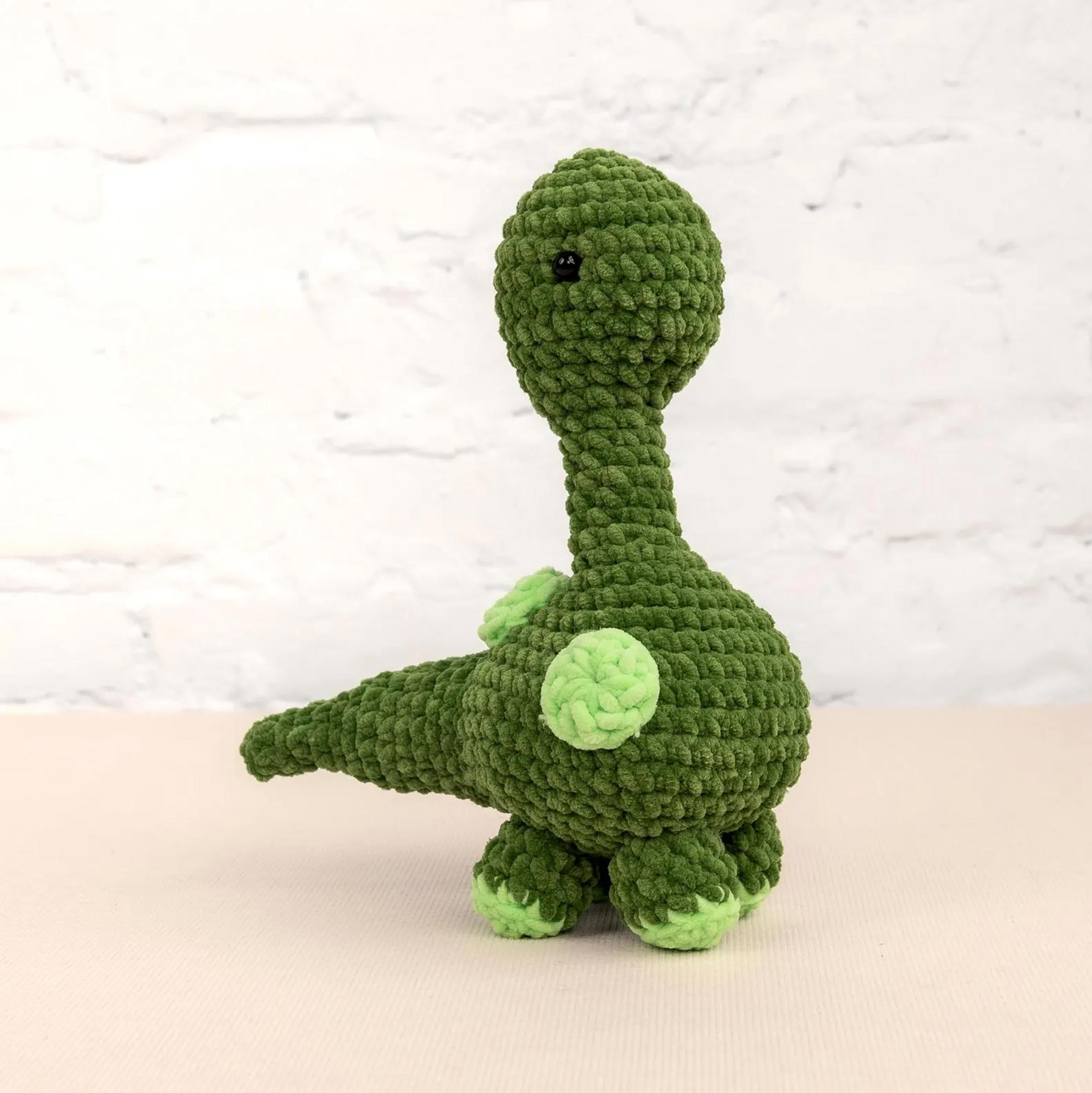 A green crocheted toy dinosaur with a round head and two green spots on its back is sitting on a white surface against a white brick wall background.