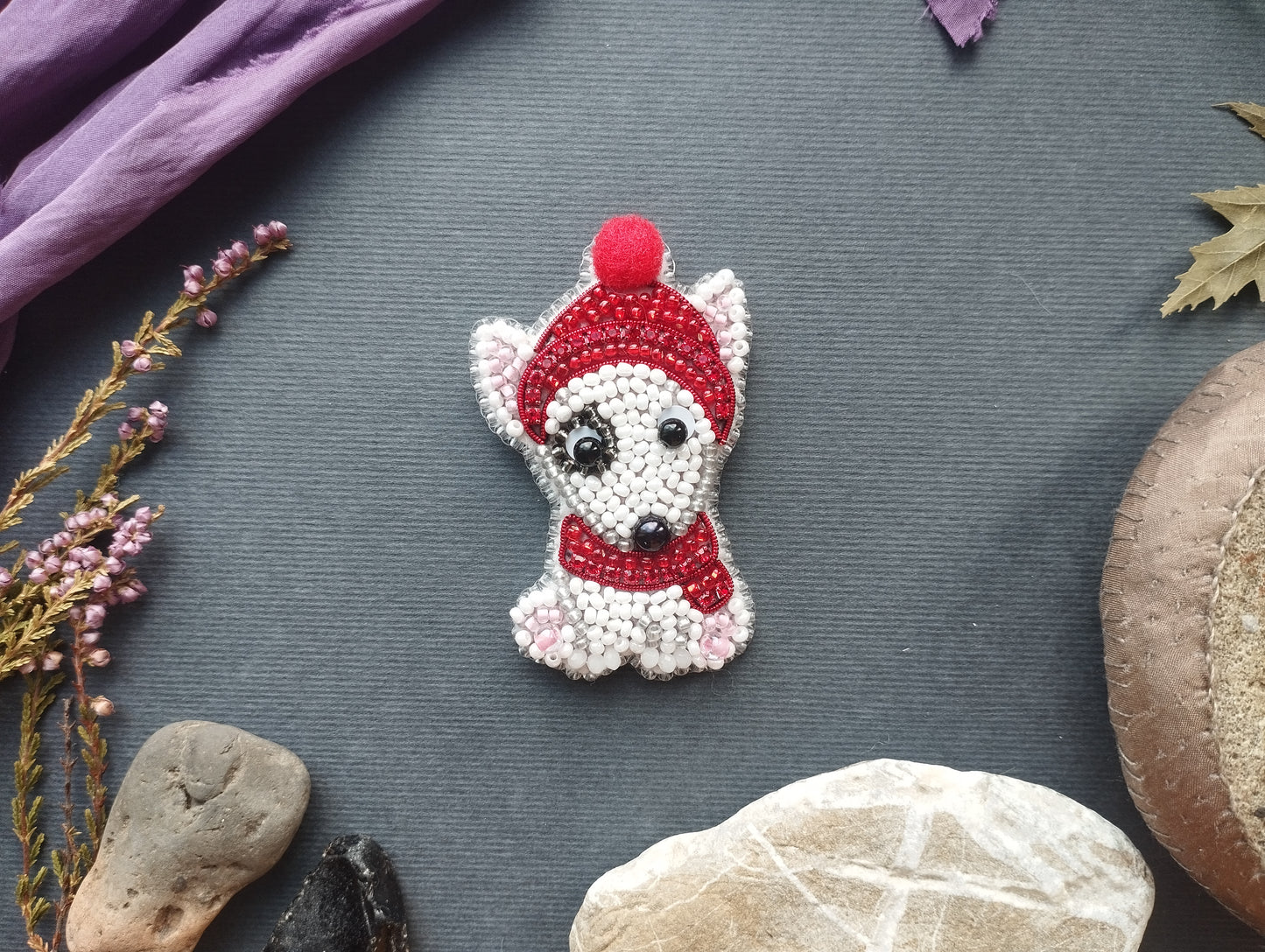 a small, intricately beaded dog brooch with a red hat and scarf, placed on a gray surface surrounded by various natural elements such as rocks, leaves, and flowers.