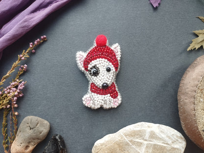 a small, intricately beaded dog brooch with a red hat and scarf, placed on a gray surface surrounded by various natural elements such as rocks, leaves, and flowers.