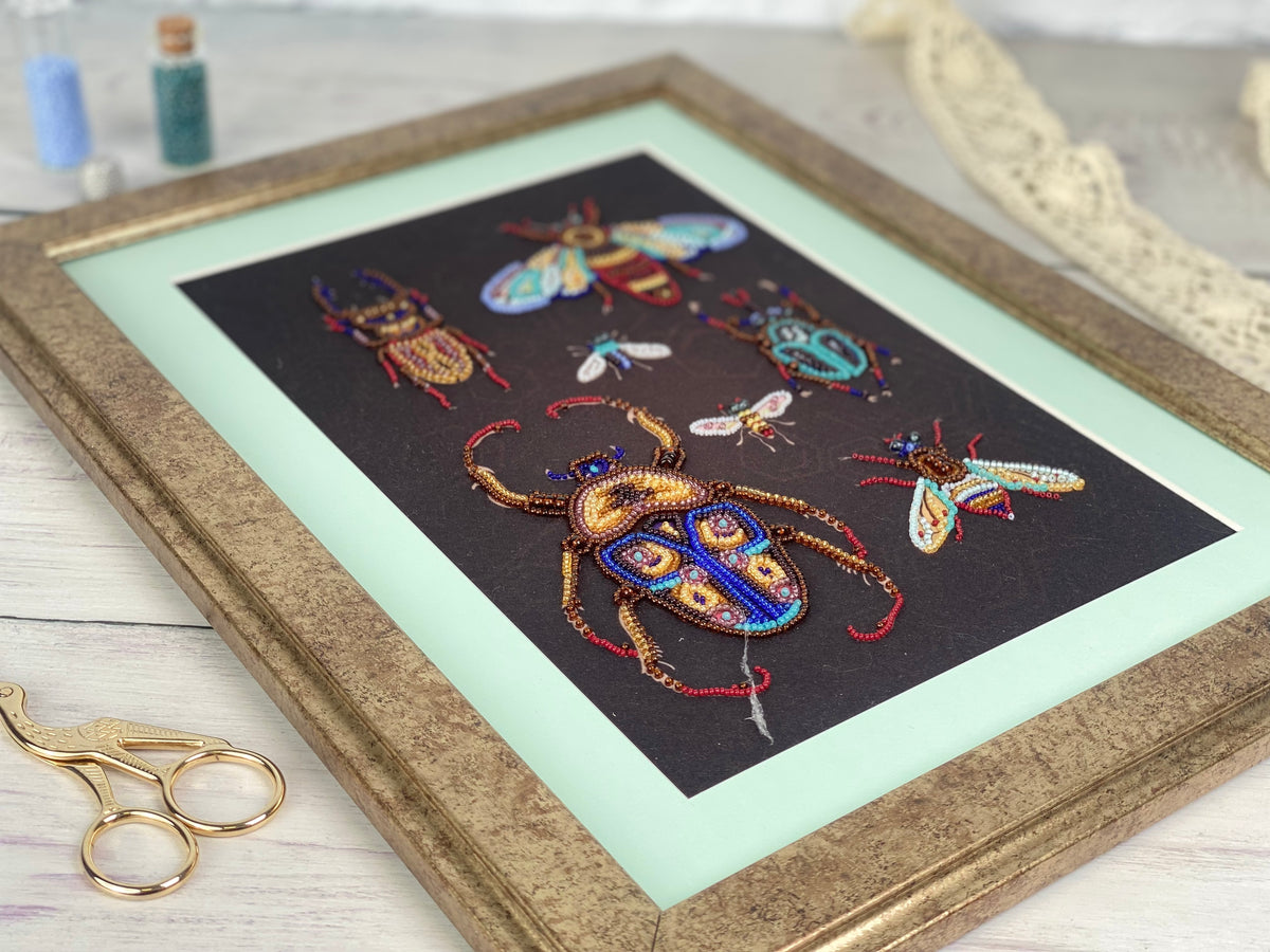 a framed artwork depicting a variety of colorful insects, including a large dragonfly and several smaller insects, all intricately embroidered or beaded.