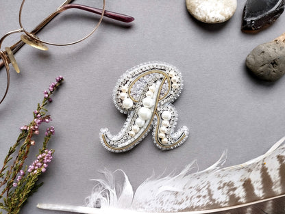 a close-up view of a decorative letter "R" made of pearls and jewels, surrounded by various objects such as glasses, feathers, and rocks.