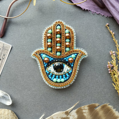 a handcrafted piece of jewelry, a hamsa charm, adorned with various colorful beads and gemstones. The charm is placed on a gray surface, surrounded by other objects such as feathers, crystals, and a card.