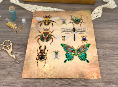 a collection of various insects, including a bee, a dragonfly, and a butterfly, all intricately detailed and adorned with jewels and beads. The insects are displayed on a piece of paper or card, which is placed on a wooden surface. In addition to the insects, there are some small objects, such as a bottle and a pair of scissors, scattered around the scene.
