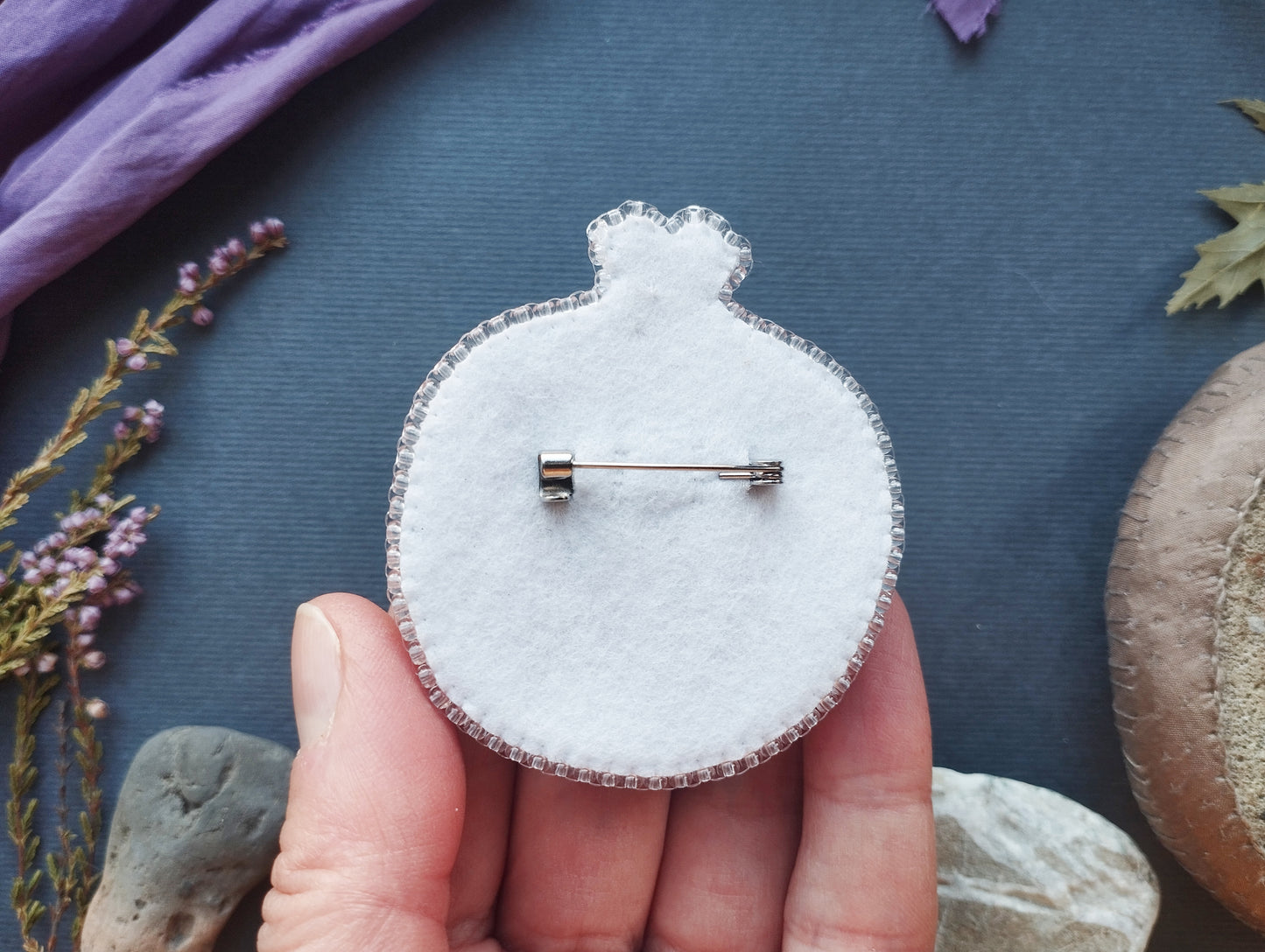 A hand holding a white felt object with a pin or pinwheel-like design on it, surrounded by various natural elements such as flowers, rocks, and leaves.