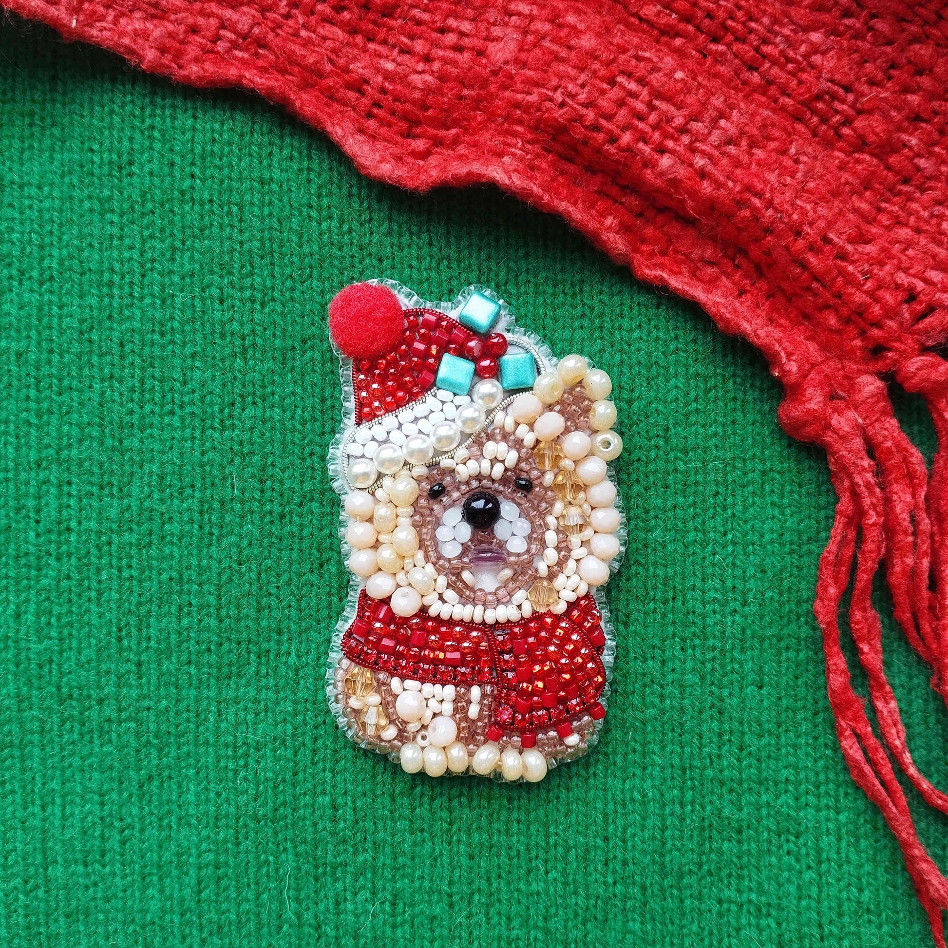 A beaded Christmas ornament in the shape of a dog wearing a Santa hat and scarf, placed on a green background with a red fringe.
