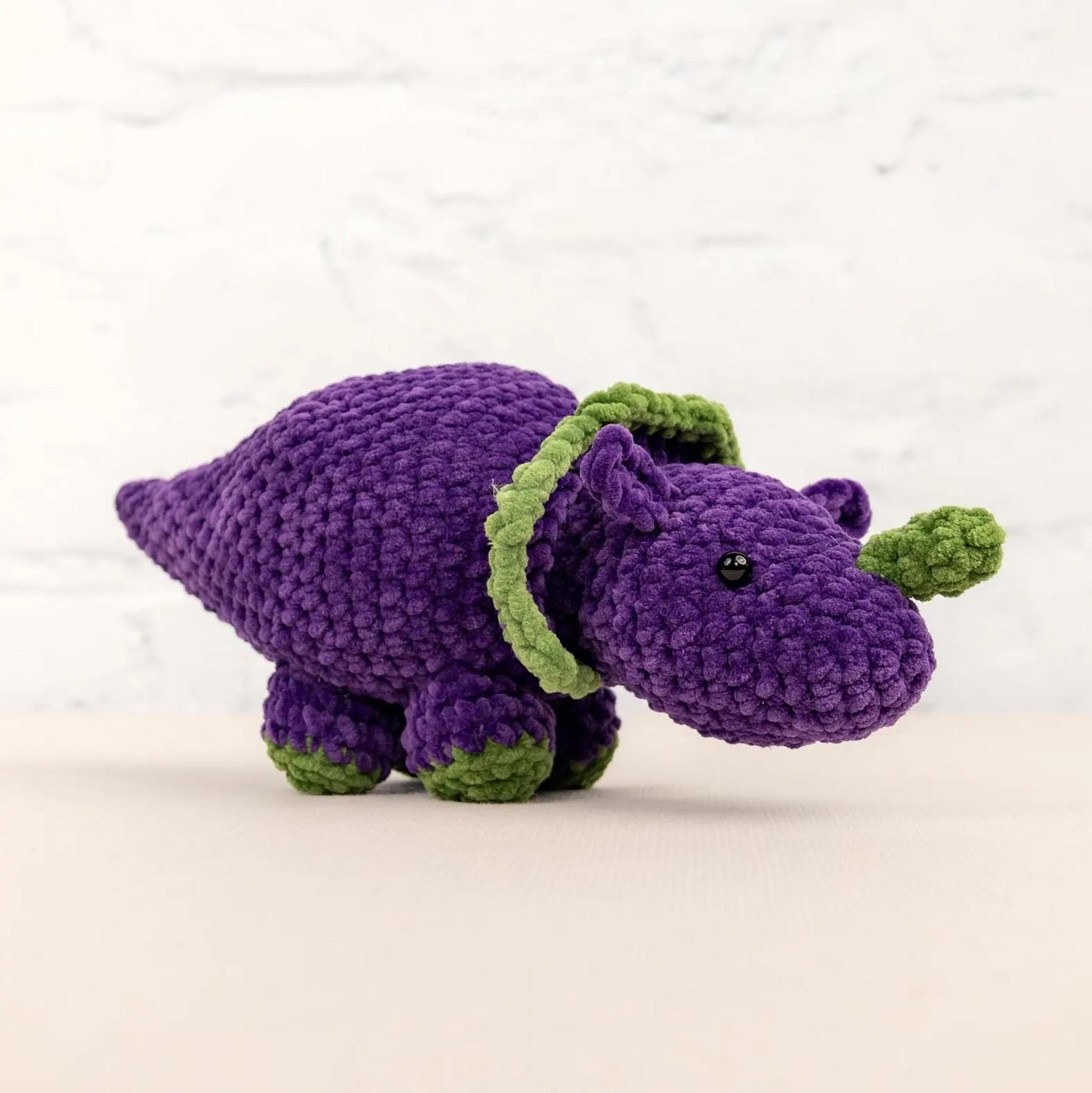 A purple crocheted toy dinosaur with green accents is sitting on a white surface against a white background.
