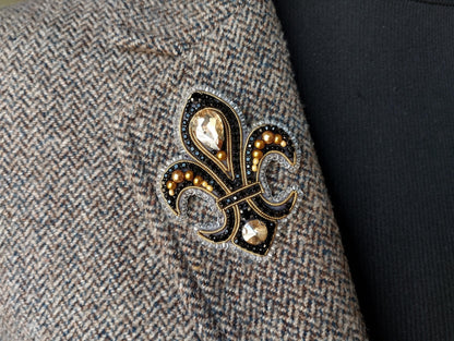 A close-up view of a grey tweed jacket with a decorative fleur-de-lis brooch featuring gold and black embellishments.