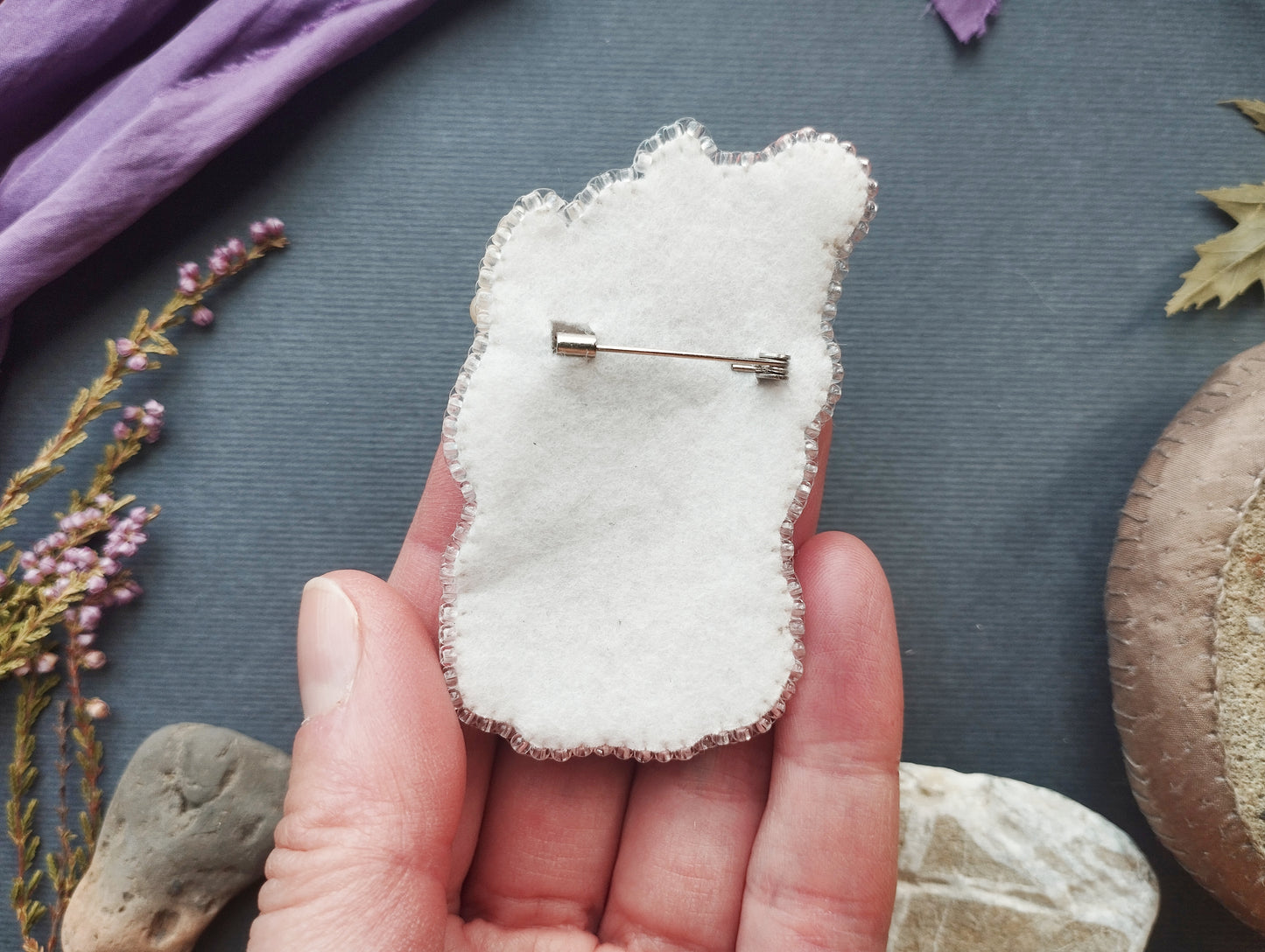 A hand holding a white fabric pin with a silver pinback attached to it, surrounded by various natural elements such as leaves, rocks, and flowers.