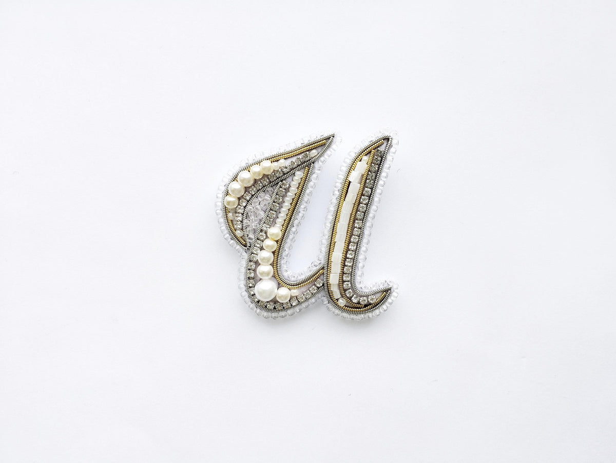 A decorative letter 'U' made of beads, pearls, and crystals against a white background.