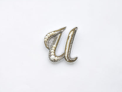 A decorative letter 'U' made of beads, pearls, and crystals against a white background.