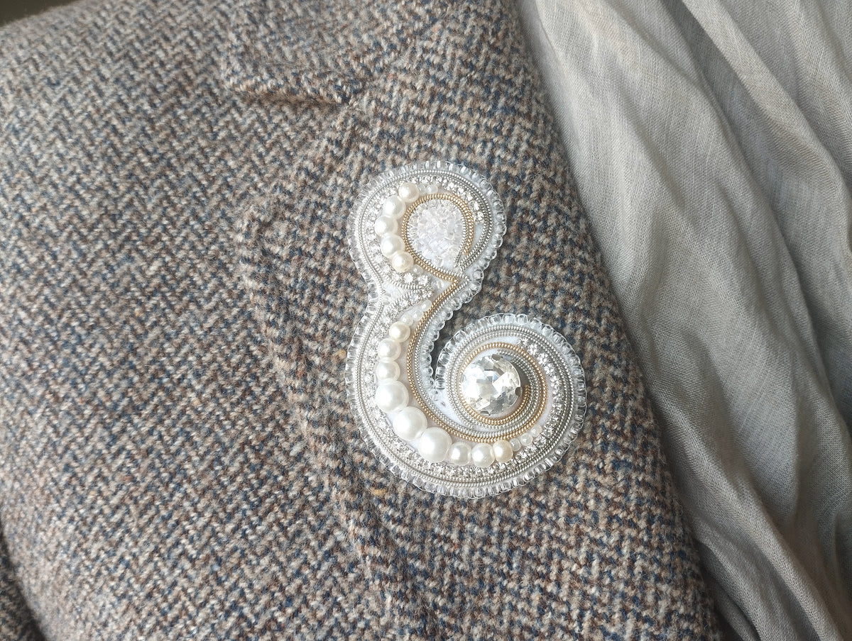 A close-up view of a grey tweed jacket with a decorative embroidered letter 'E' on the lapel.