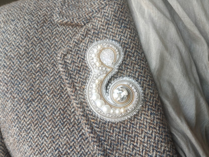 A close-up view of a grey tweed jacket with a decorative embroidered letter 'E' on the lapel.