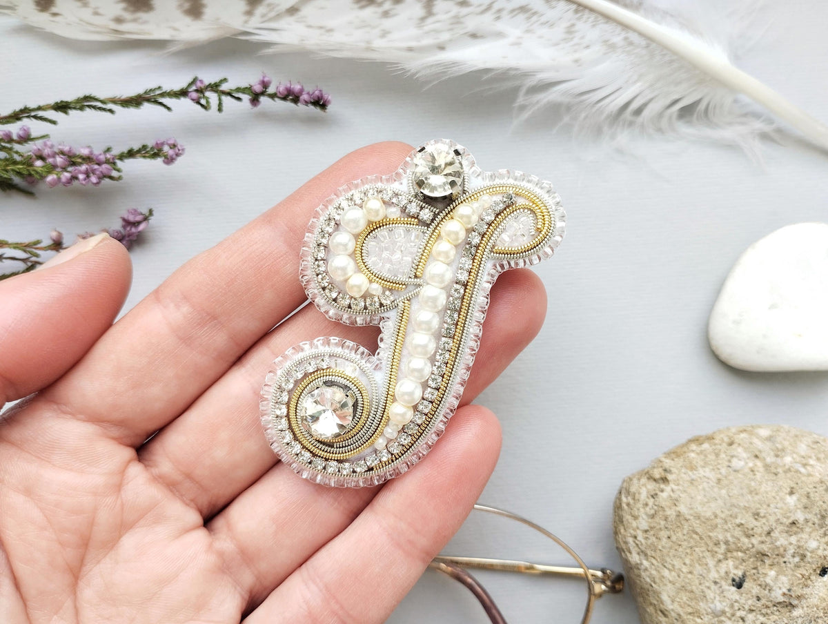 A hand holding a decorative letter "J" made of beads and pearls, with a gemstone at the top.