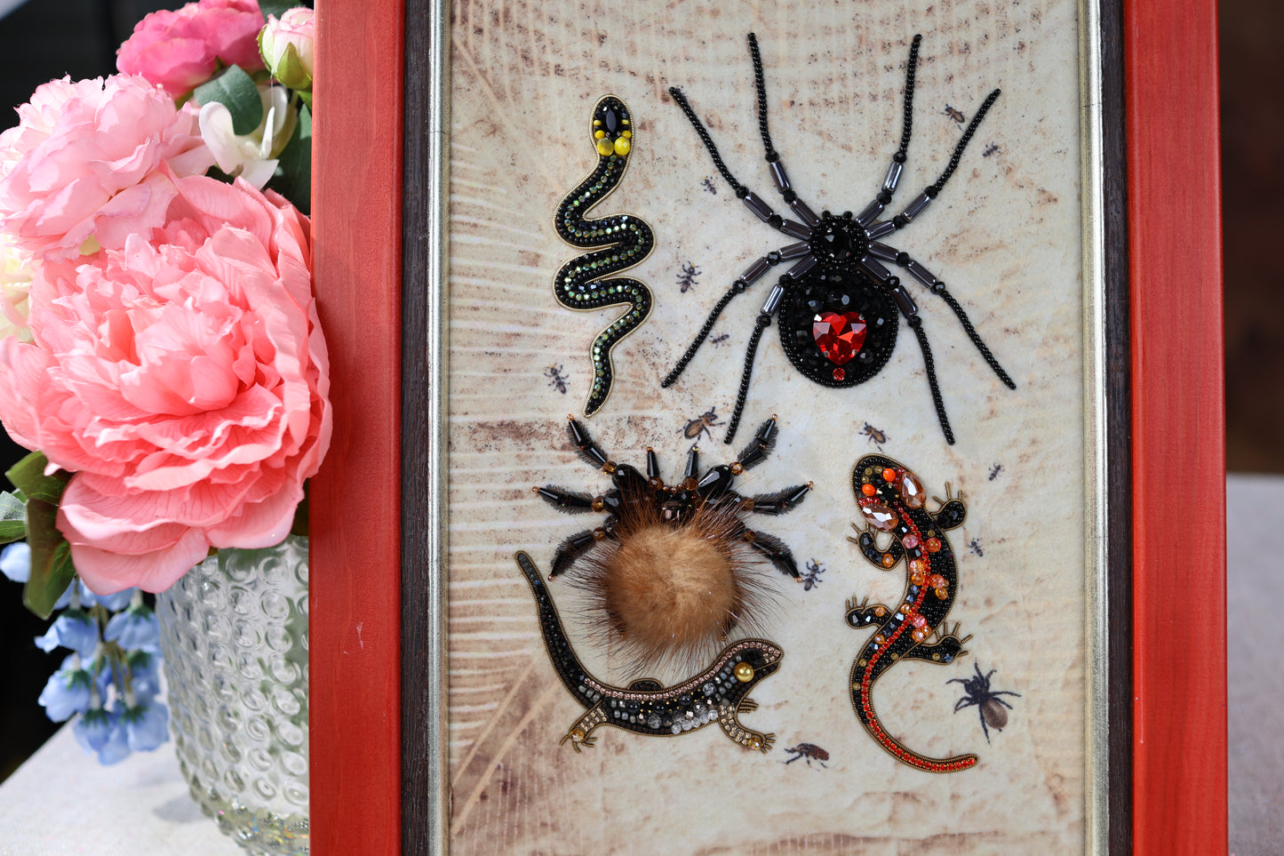 a close-up of a framed collage of various insects, including a spider, a lizard, and a bee, all adorned with beaded or embroidered details. The insects are displayed against a textured background, creating a visually striking and detailed composition.