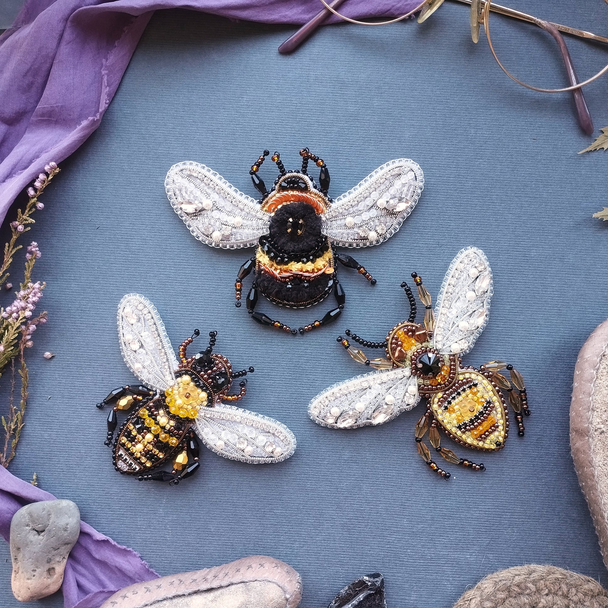 three intricately designed beaded bee brooches arranged on a blue background.