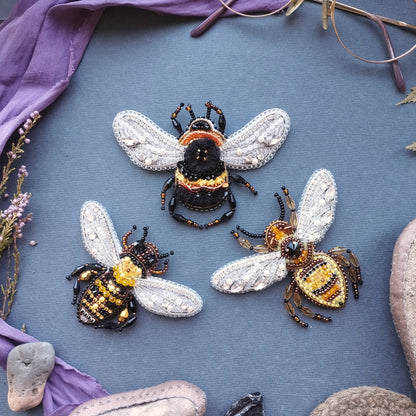 three intricately designed beaded bee brooches arranged on a blue background.