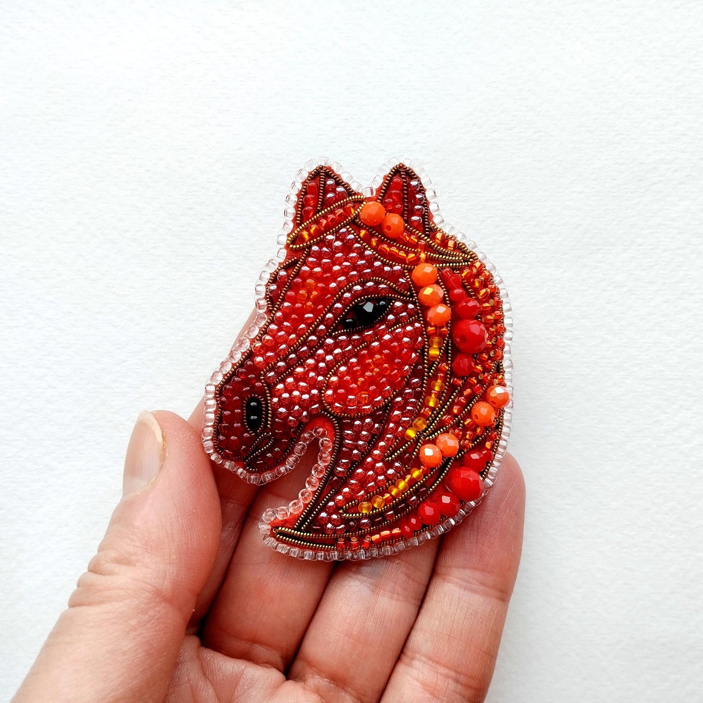 A hand holding a decorative piece of red and orange beaded embroidered in the shape of a horse's head.