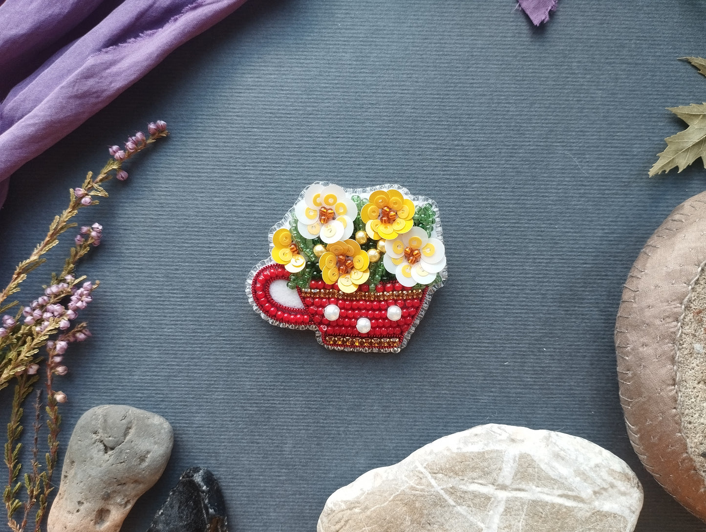 a red and white beaded cup-shaped brooch with yellow and white flowers, placed on a gray surface surrounded by various natural elements such as rocks, leaves, and flowers.