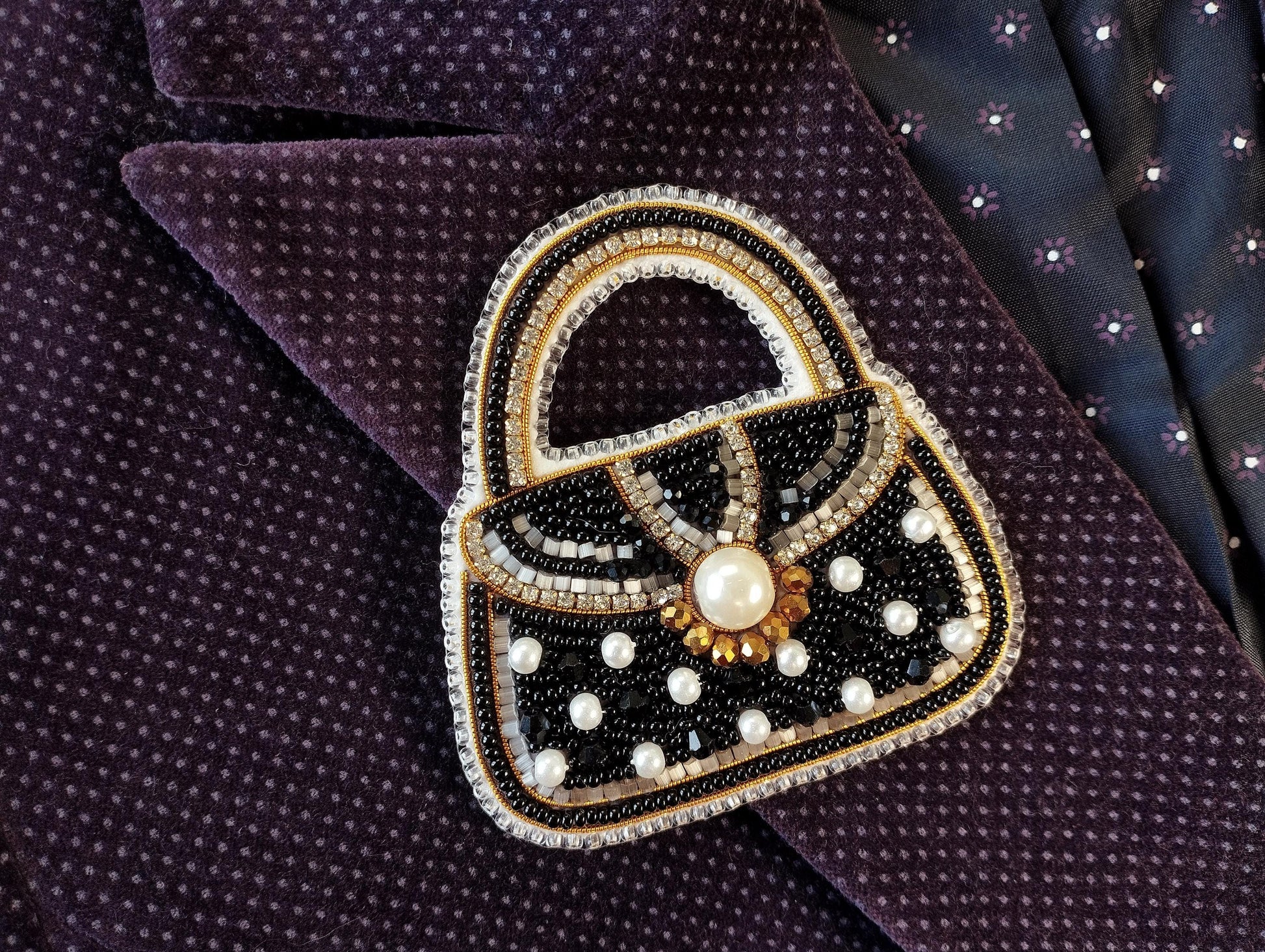 A hand-made purse brooch with a black and gold design, featuring a pearl at the center and beaded embellishments, is displayed on a purple fabric with a polka dot pattern.