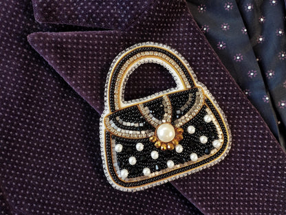 A hand-made purse brooch with a black and gold design, featuring a pearl at the center and beaded embellishments, is displayed on a purple fabric with a polka dot pattern.