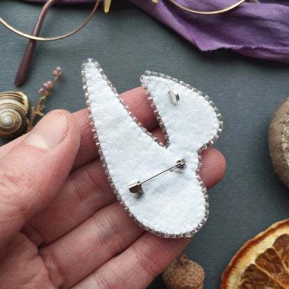 A hand holding a white felt pin with a silver pin attached to it, surrounded by various objects such as a pair of glasses, a purple fabric, and dried fruit.