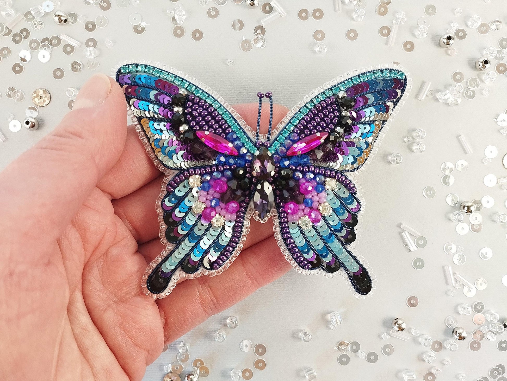 A hand is holding a colorful, sequined butterfly brooch against a background of sequins and beads.