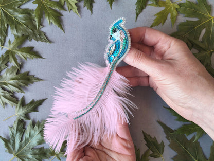 A person&#39;s hand holding a decorative feather with a blue and green beaded design, surrounded by green leaves.