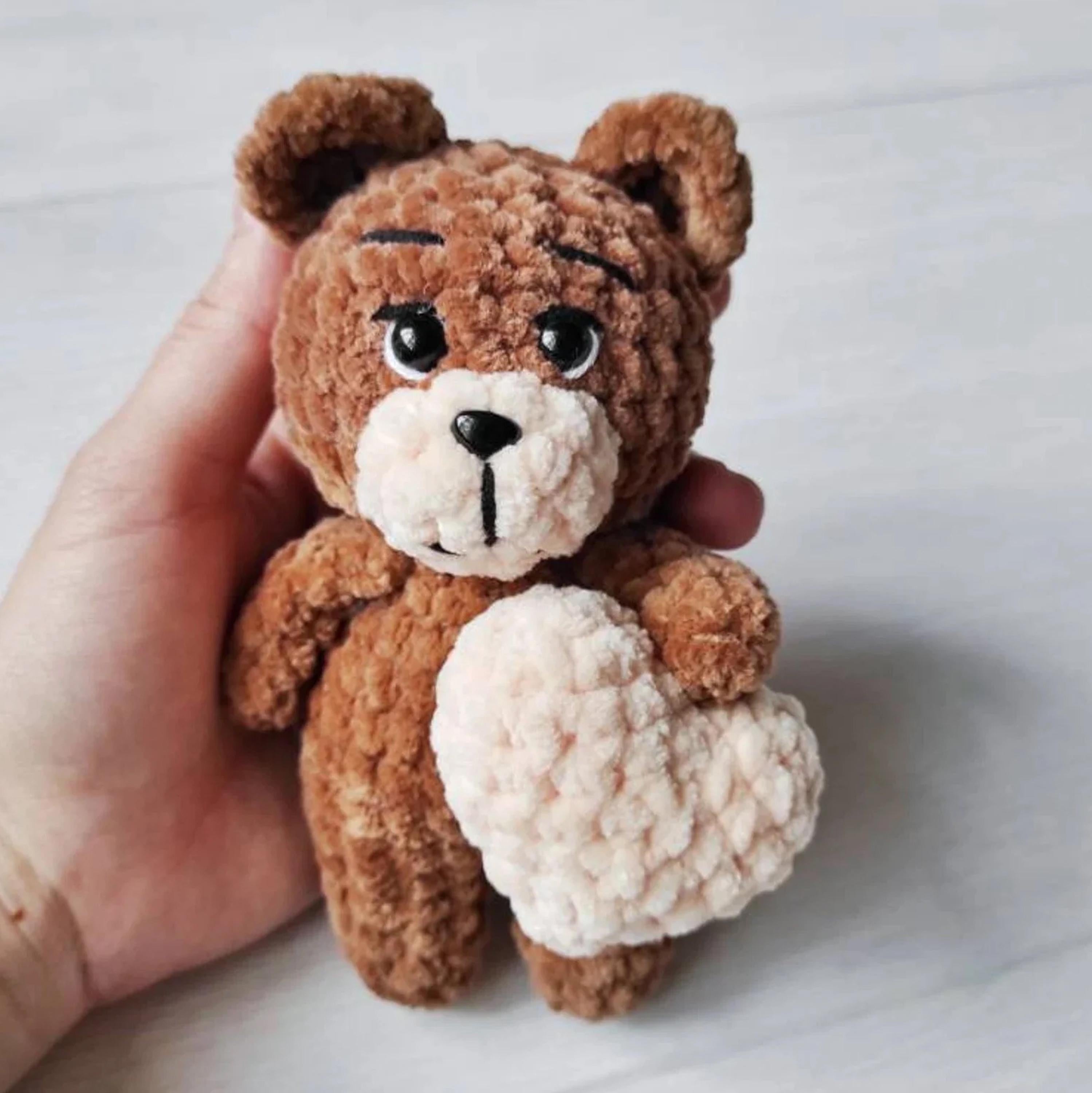 A hand is holding a brown teddy bear with a heart-shaped object in its paws.