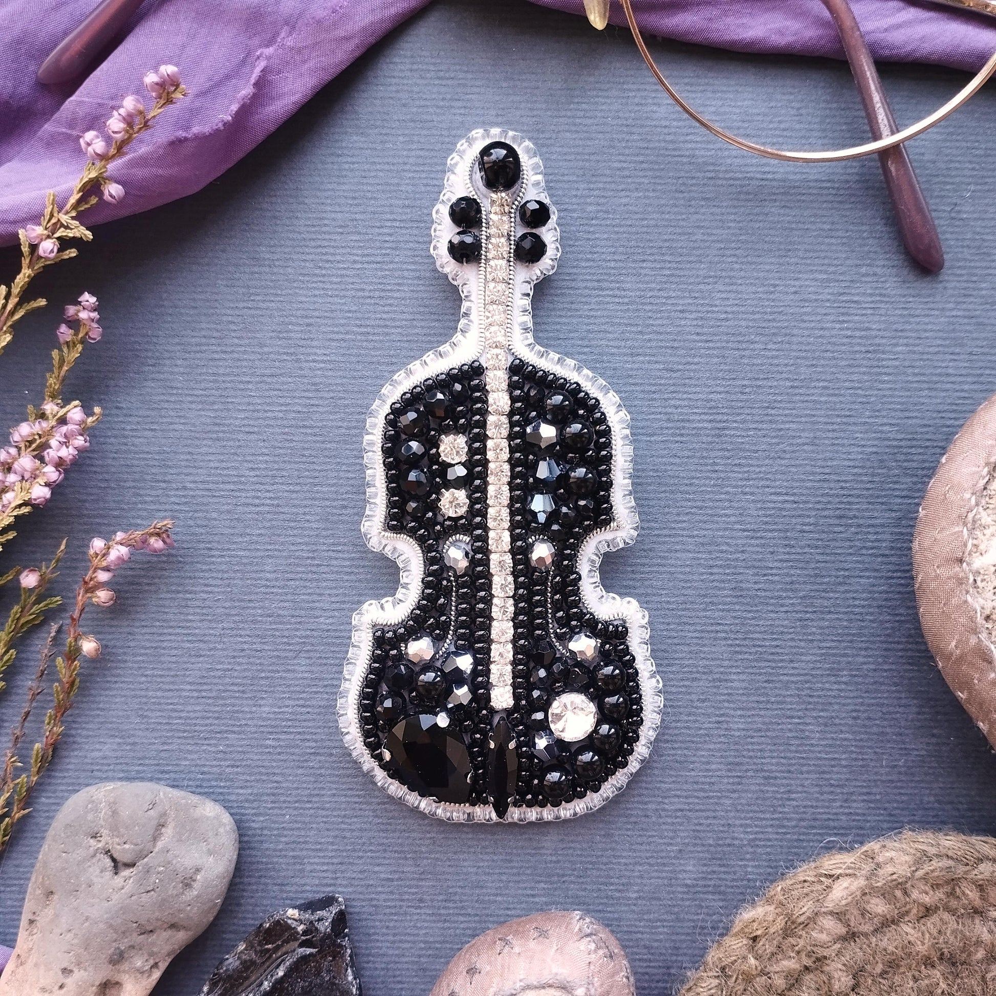 a black and white beaded violin brooch, surrounded by various objects such as a pair of glasses, a flower, and a rock.