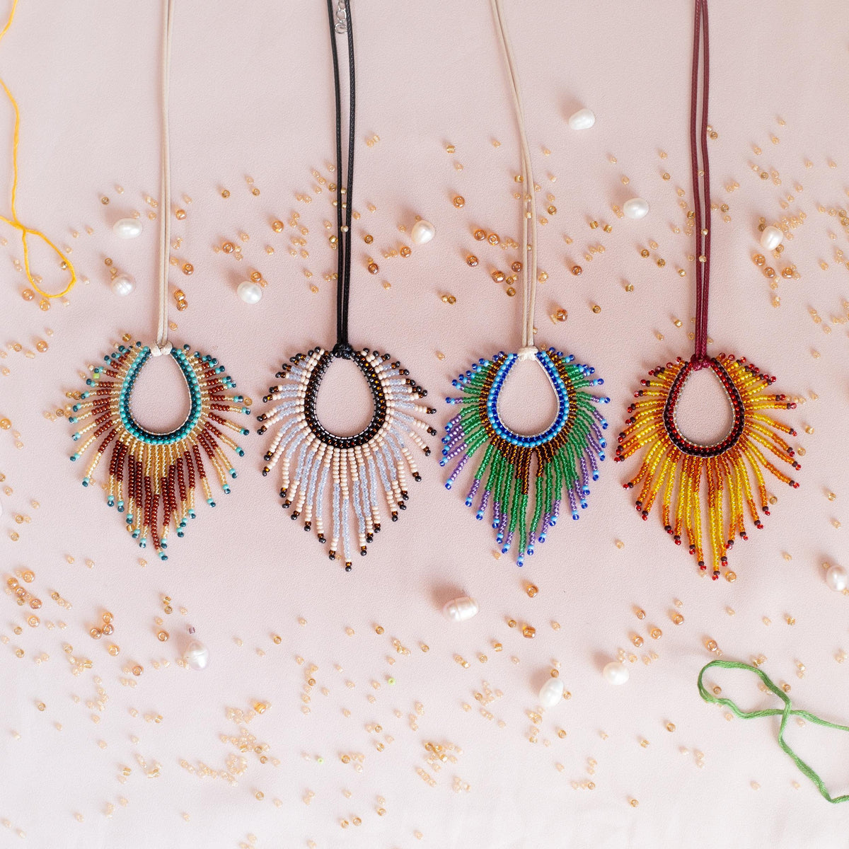 four colorful beaded necklaces with feather-like designs, each with a unique color and pattern, arranged on a pink background with scattered beads.