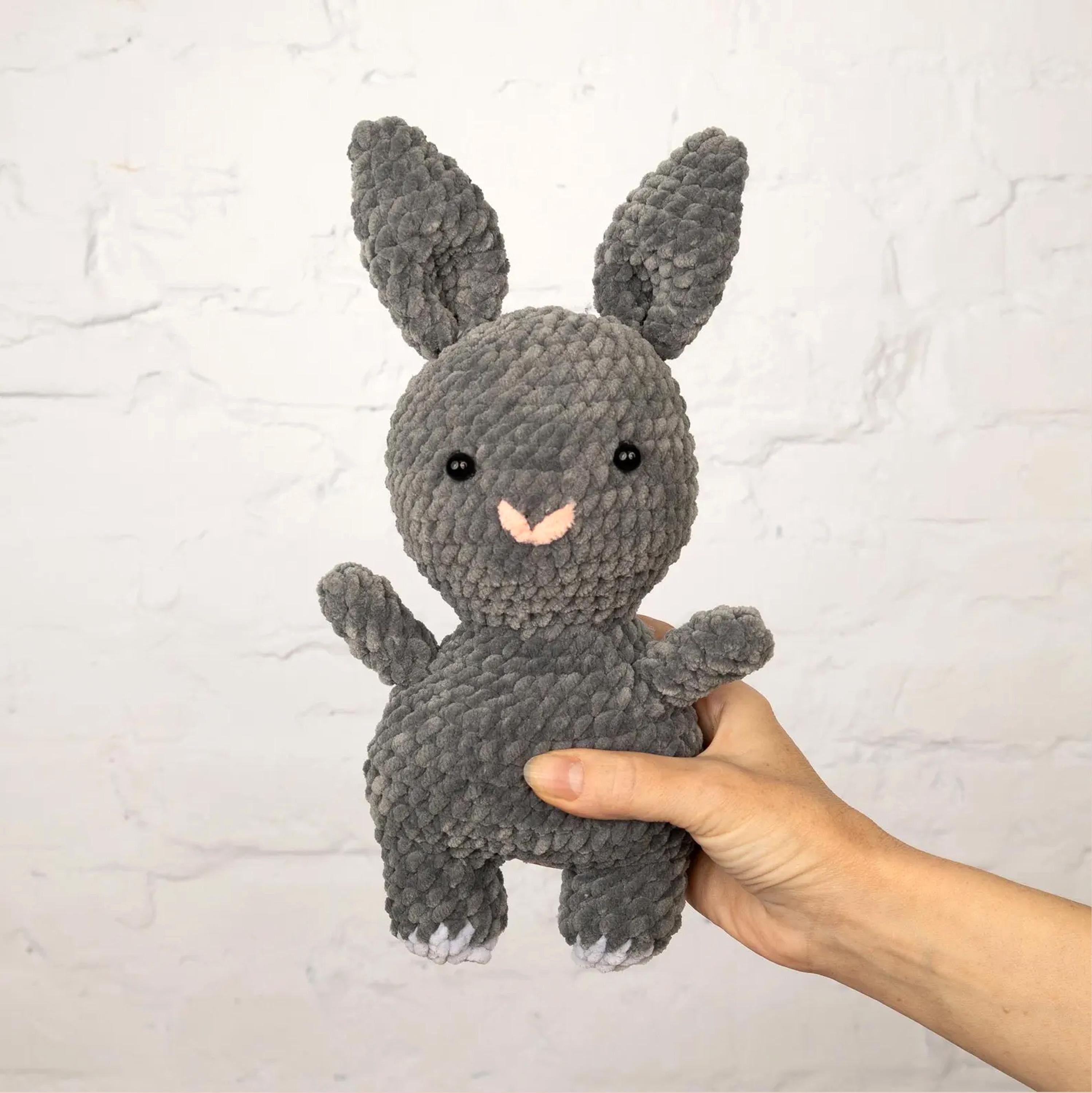 A hand is holding a gray, crocheted stuffed rabbit toy against a white background.