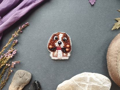 a small, intricately designed dog brooch made of beads and surrounded by various natural elements such as rocks, leaves, and flowers.