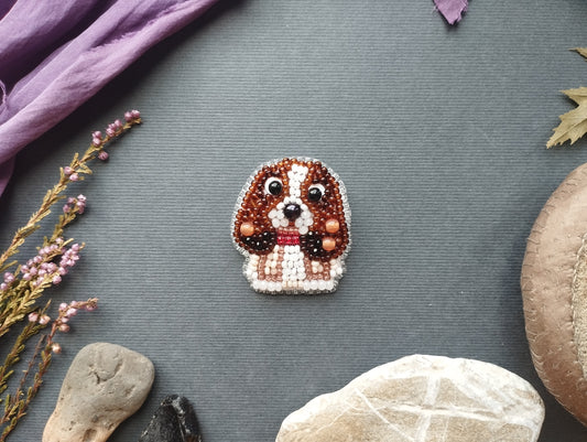 a small, intricately designed dog brooch made of beads and surrounded by various natural elements such as rocks, leaves, and flowers.