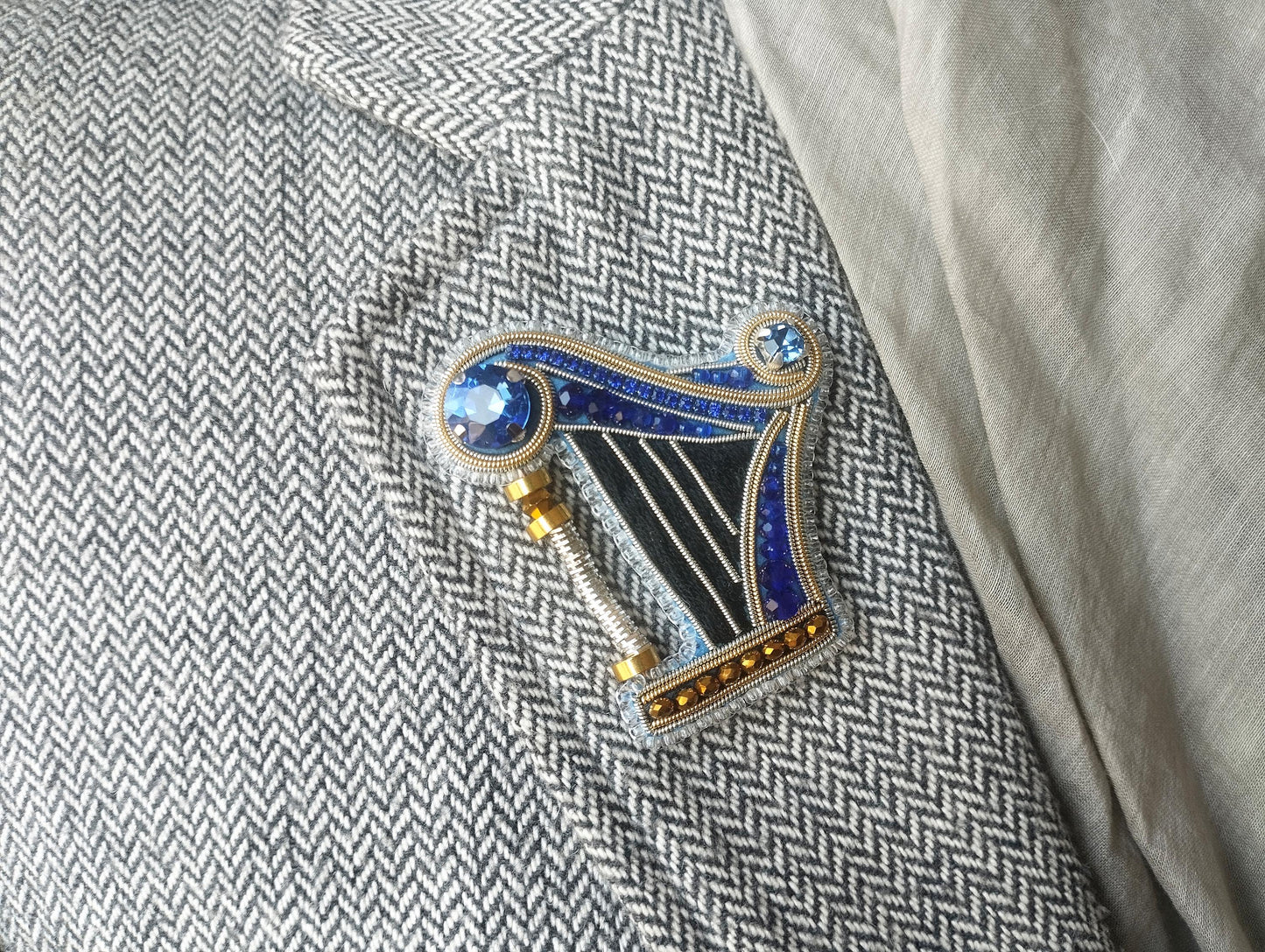 A decorative brooch in the shape of a harp, adorned with blue and gold gemstones, is displayed on a gray and white checkered fabric.