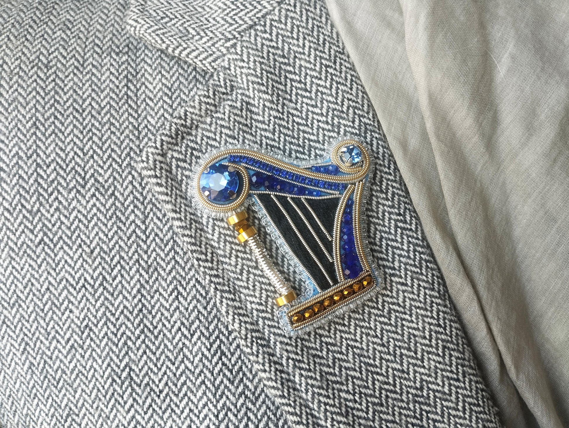 A decorative brooch in the shape of a harp, adorned with blue and gold gemstones, is displayed on a gray and white checkered fabric.