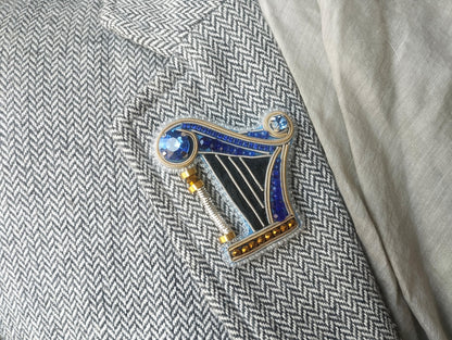 A decorative brooch in the shape of a harp, adorned with blue and gold gemstones, is displayed on a gray and white checkered fabric.
