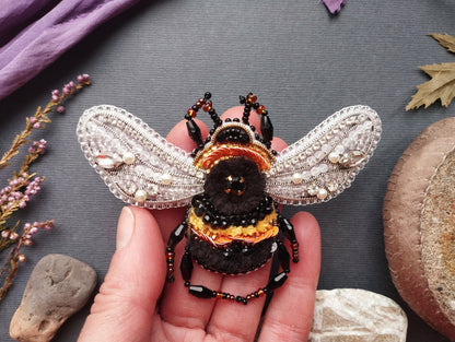 A hand is holding a decorative beaded insect brooch, which is a detailed and intricate piece of jewelry. The hand is positioned over a variety of objects, including rocks, leaves, and flowers, creating a natural and artistic composition.