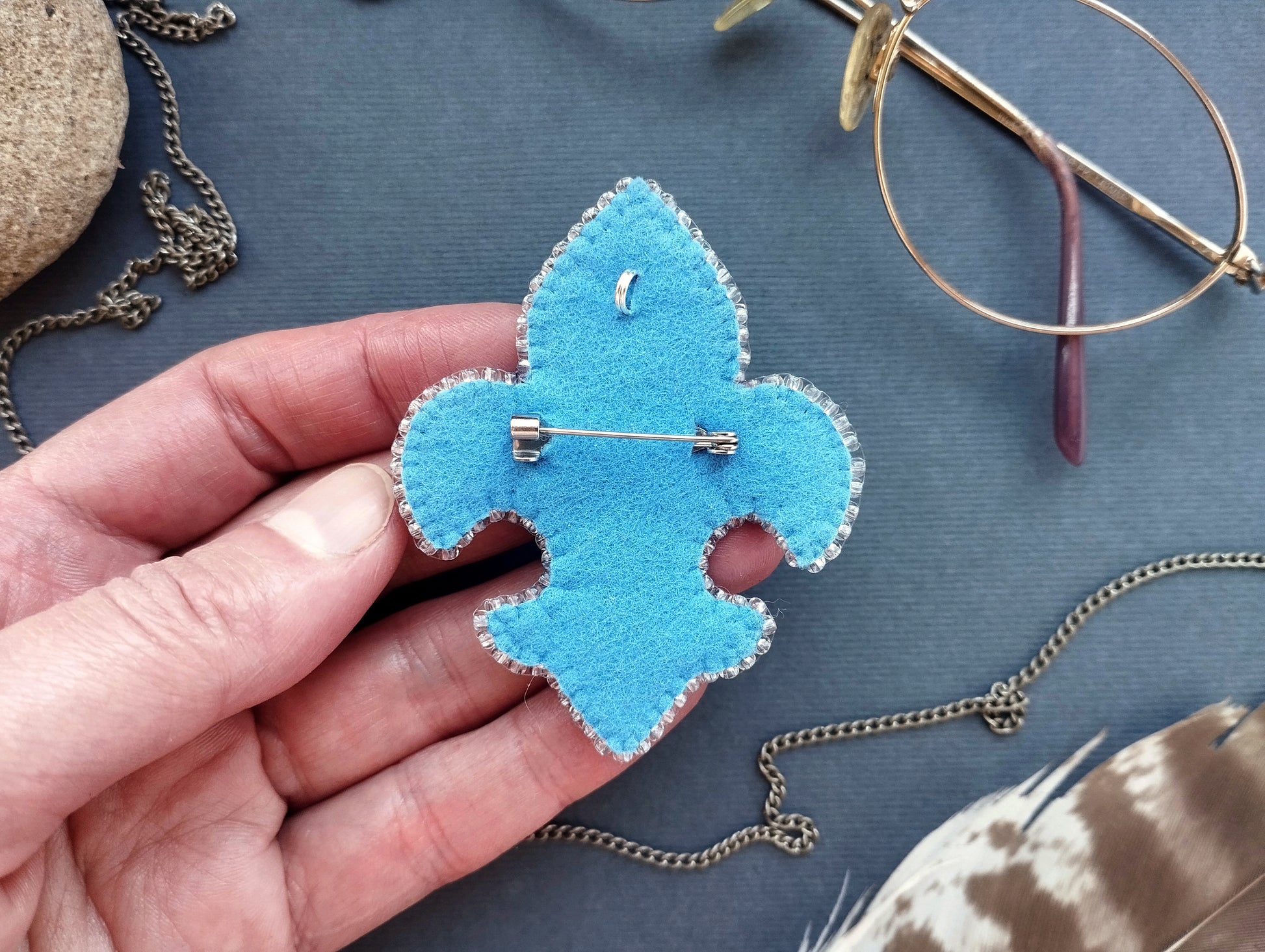 A hand is holding a blue felt badge with a pin on it, surrounded by various accessories such as glasses, a chain, and feathers.