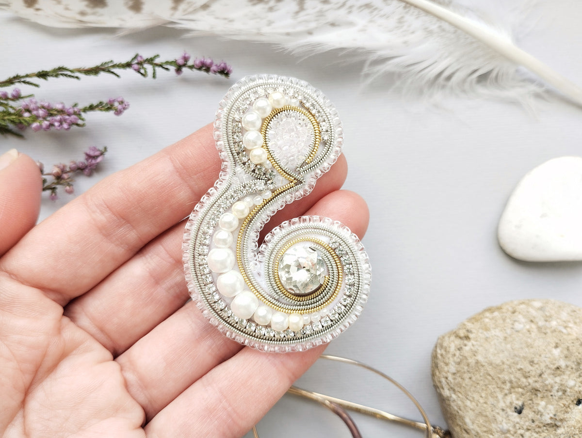 A hand holding a decorative piece of jewelry, which appears to be a brooch or pin, with intricate beading and pearl details. The background includes various natural elements such as feathers and rocks, creating a serene and natural setting.