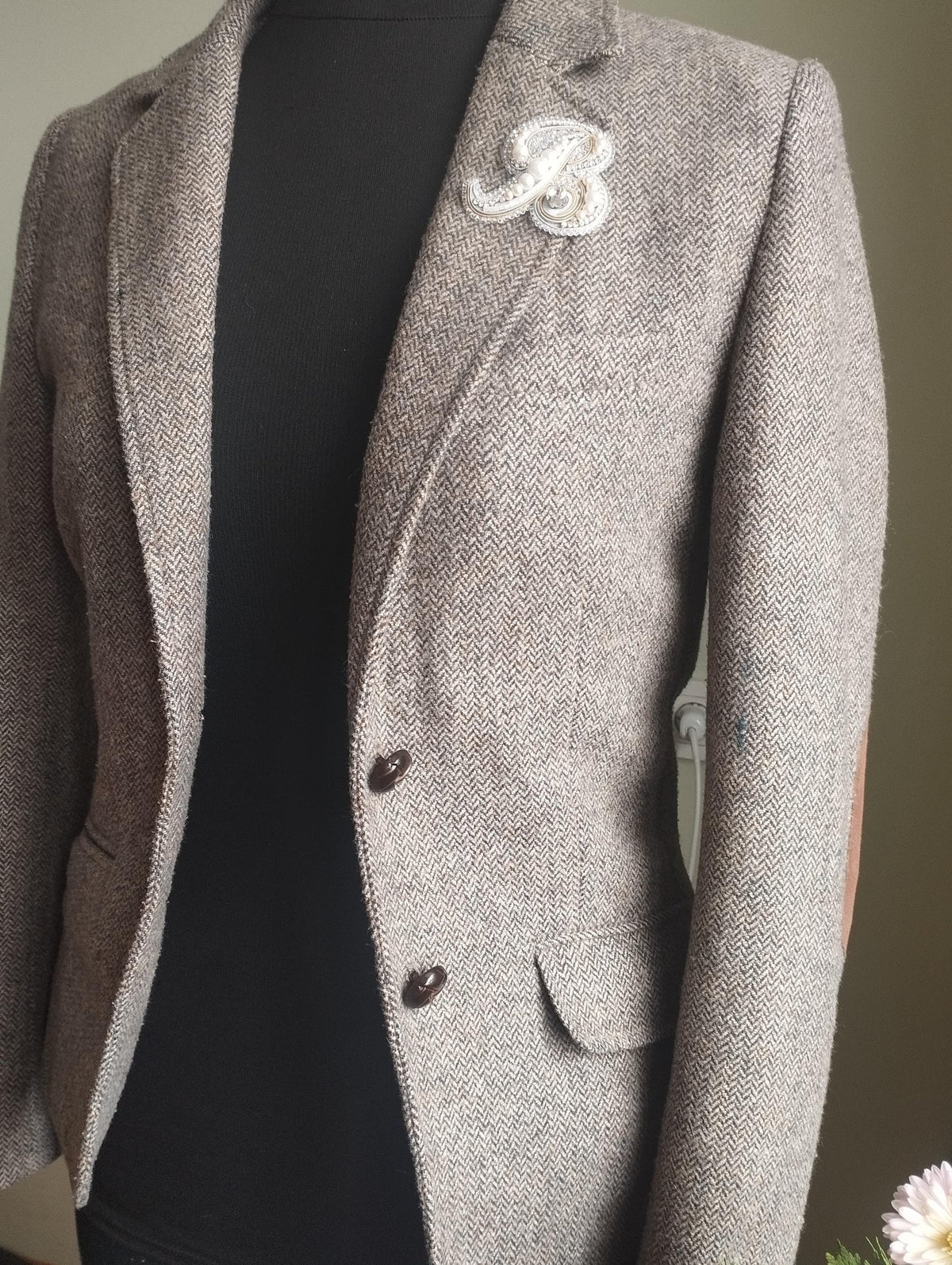 A gray tweed blazer with a white "B" brooch on the left breast is displayed on a mannequin.