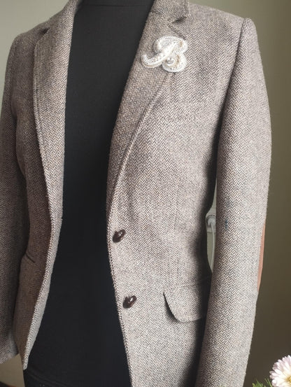 A gray tweed blazer with a white "B" brooch on the left breast is displayed on a mannequin.