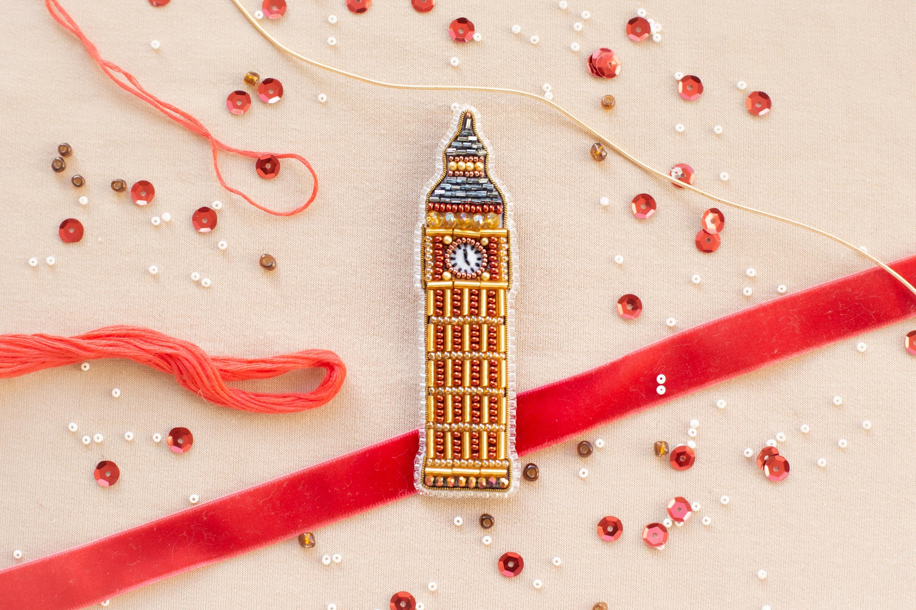 a close-up view of a small, intricately designed embroidered object resembling Big Ben, surrounded by various craft supplies such as beads, string, and a red ribbon.