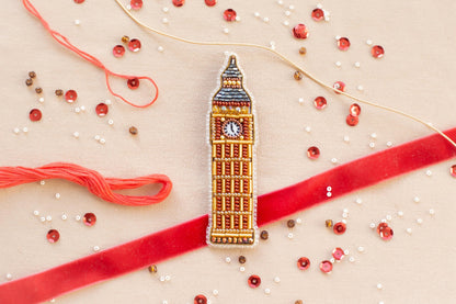 a close-up view of a small, intricately designed embroidered object resembling Big Ben, surrounded by various craft supplies such as beads, string, and a red ribbon.