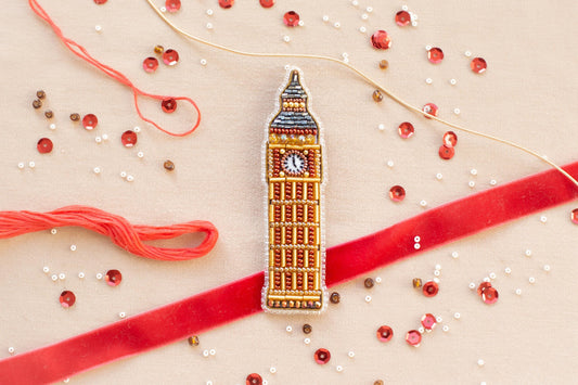 a close-up view of a small, intricately designed embroidered object resembling Big Ben, surrounded by various craft supplies such as beads, string, and a red ribbon.