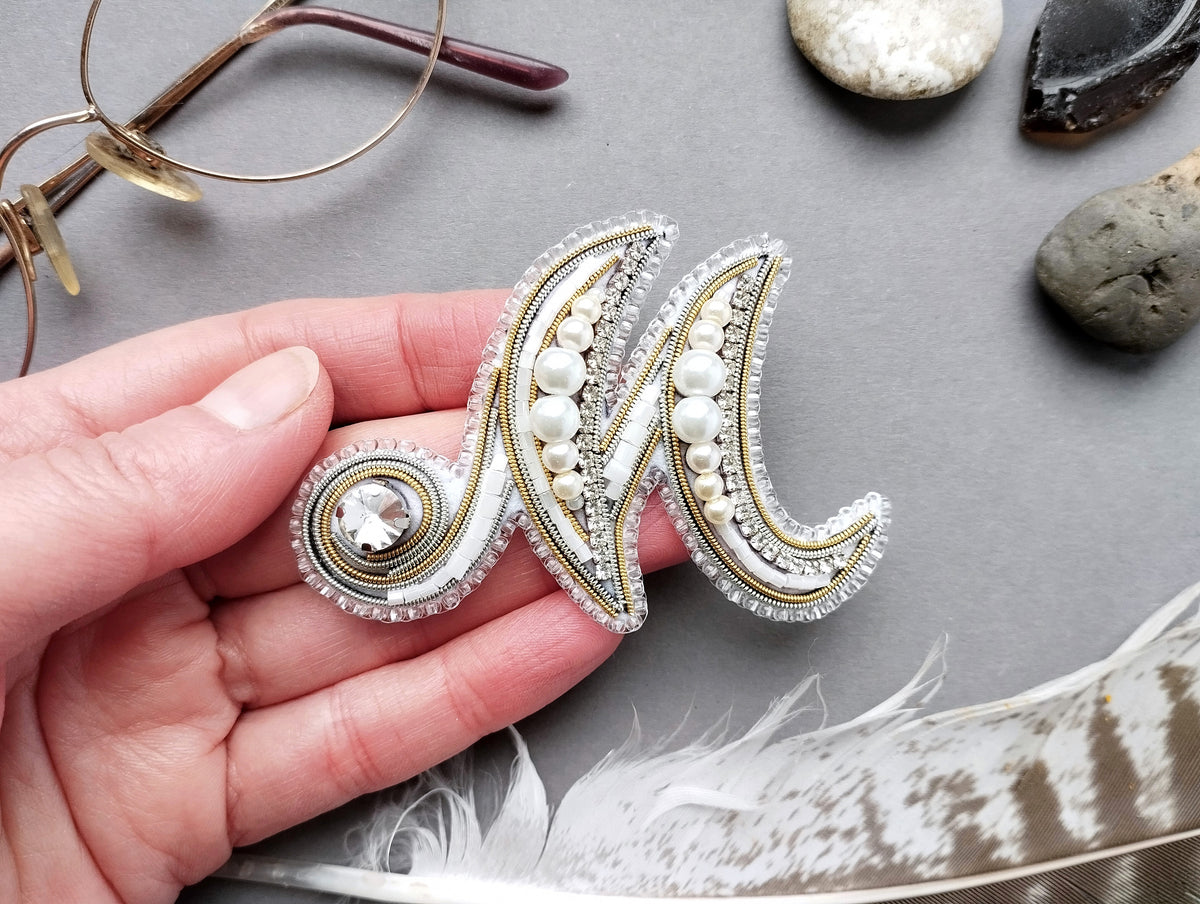 A hand holding a decorative monogram brooch with pearls and gemstones, surrounded by various objects such as glasses, rocks, and feathers.