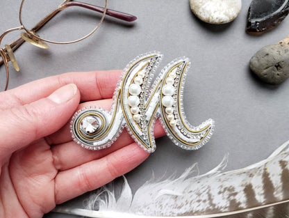 A hand holding a decorative monogram brooch with pearls and gemstones, surrounded by various objects such as glasses, rocks, and feathers.