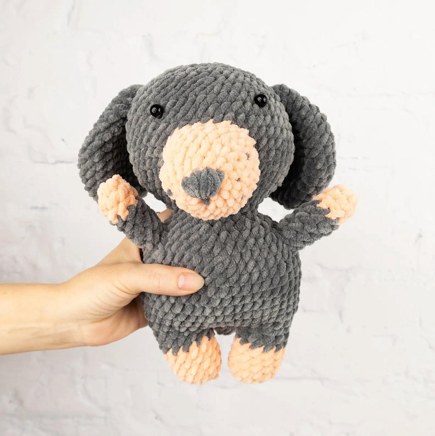 A hand is holding a gray and peach-colored stuffed animal, which appears to be a small dog or puppy.